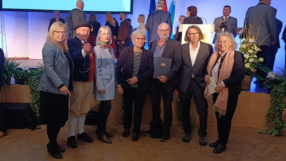 Eine Gruppe von sieben Personen steht lächelnd für ein Foto auf einer Bühne. Ein Mann in traditioneller Tracht und ein Mann mit einer Urkunde in den Händen befinden sich unter den Geehrten